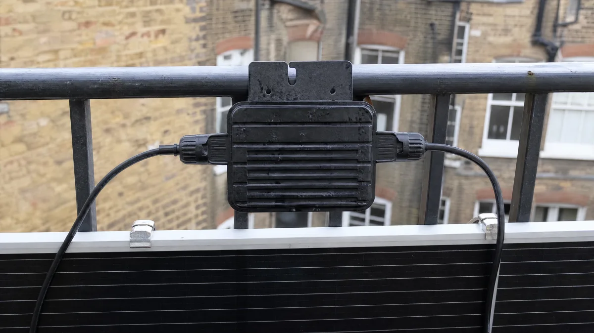 A small black weatherproof microinverter box with two cables attached, mounted behind a solar panel on a metal balcony railing.